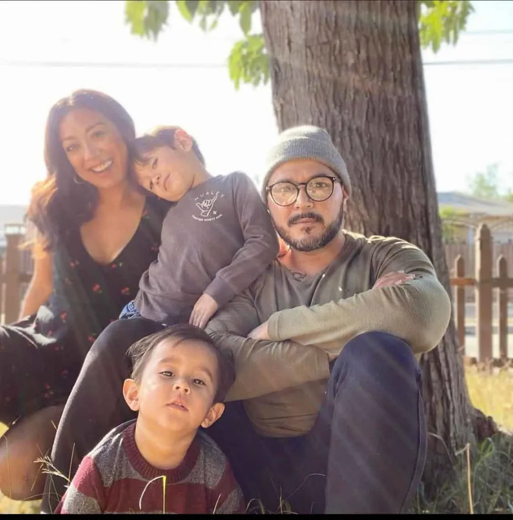Family of four seated outdoors near a tree, with sunlight filtering through green leaves.