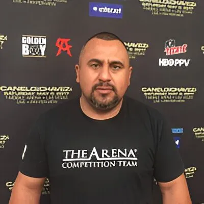 Man with short hair and beard wearing a black "The Arena Competition Team" t-shirt, standing before a black promotional backdrop.