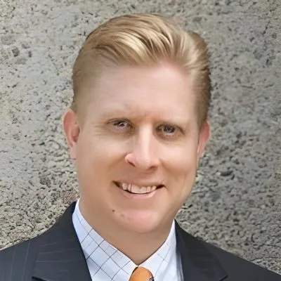 Portrait of a smiling man with blonde hair wearing a dark suit, white shirt, and orange tie.