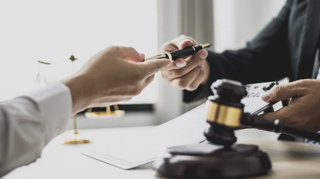 Two individuals exchanging a pen over legal documents with a judge's gavel and scales of justice in the background.