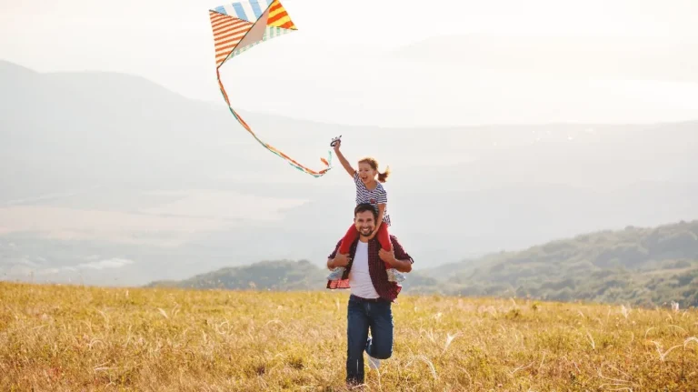 Man running in a field carrying a smiling child on shoulders flying a colorful kite.