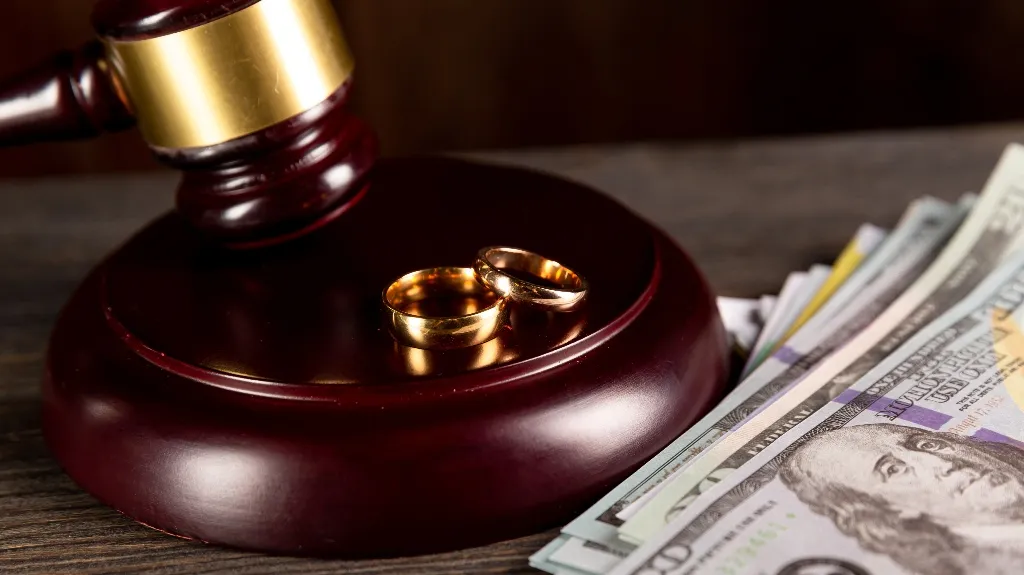 Close-up of a wooden judge's gavel with two gold wedding rings and U.S. currency on a wooden surface.