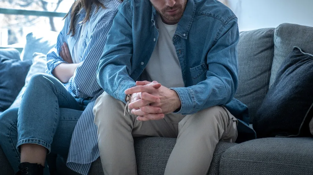 Two people sitting on a gray couch, facing away from each other, appearing upset.