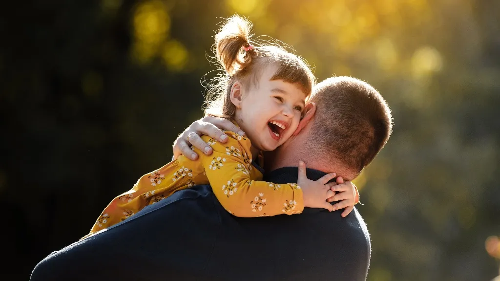 Young girl in a yellow floral dress joyfully hugging a man in a dark shirt outdoors.