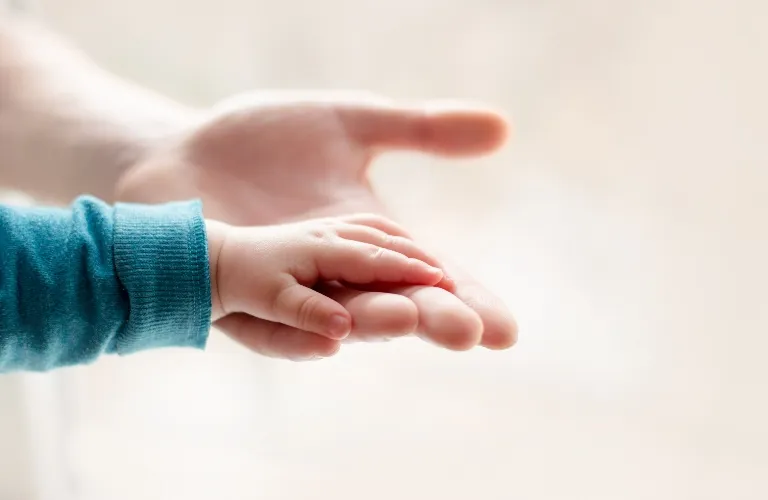 Close-up of an adult hand gently holding a baby's hand against a soft beige background.