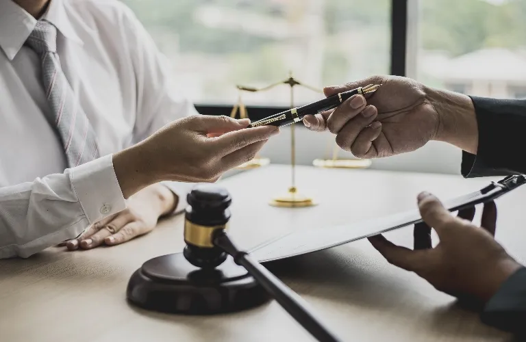 Two individuals exchanging a pen over a desk with a gavel and legal scales in the background.