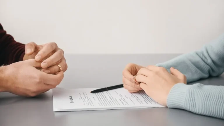Two people holding wedding rings over a divorce agreement document on a gray table.