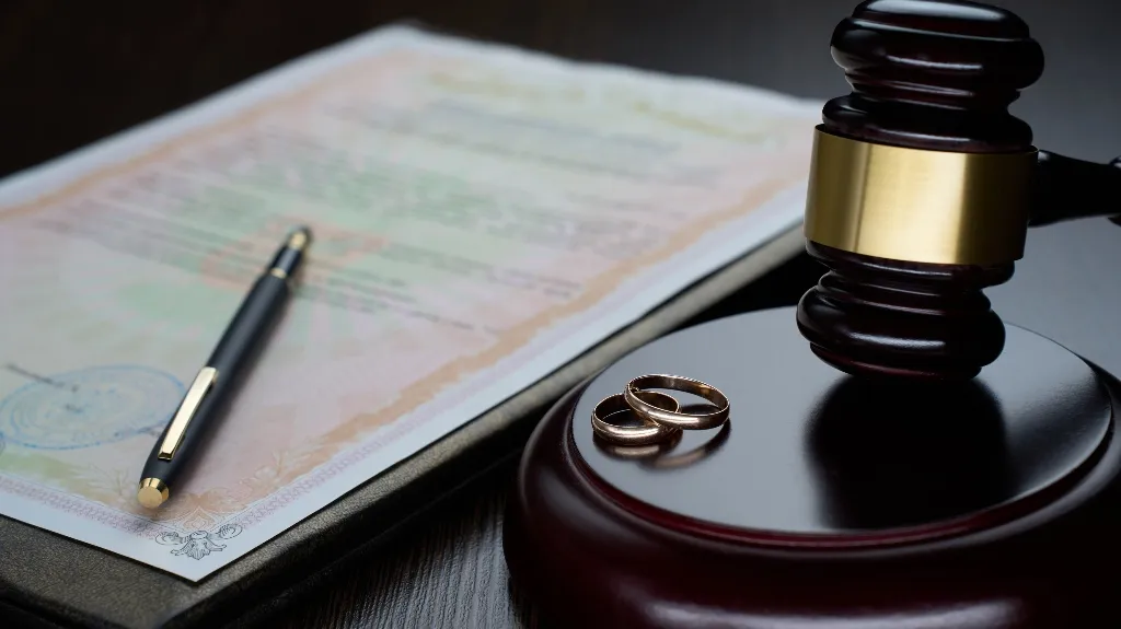 Wooden judge's gavel with two gold wedding rings and a legal document with pen on dark surface.