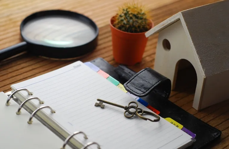 Open binder with vintage key on lined paper, small potted cactus, magnifying glass, and wooden birdhouse on wooden surface.