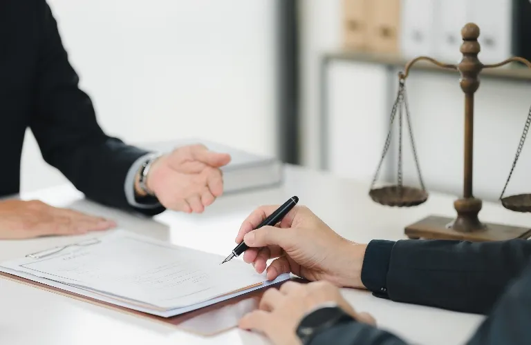 Two individuals in business attire at a desk with legal scales and a document being signed.