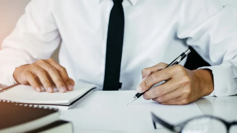 Close-up of a person in a white shirt and black tie writing on paper with a pen at a desk.