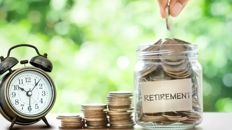 Hand placing a coin into a clear jar labeled "RETIREMENT" next to stacked coins and a vintage alarm clock.