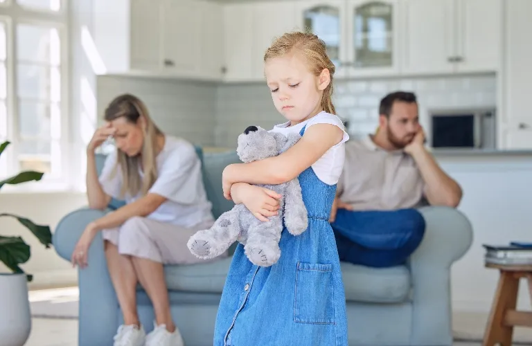 Young girl in blue dress hugging stuffed bear, parents sitting upset on couch behind.