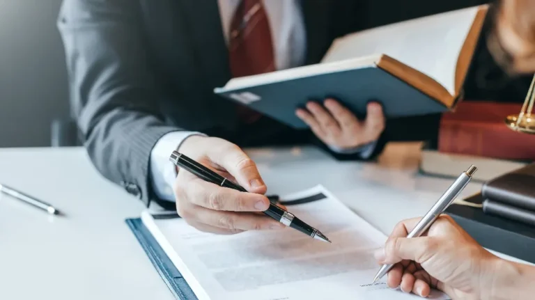 Two professionals in suits reviewing and signing documents at a desk with books.