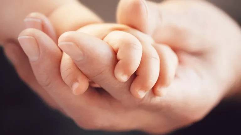 Close-up of an adult hand gently holding a newborn baby's hand against a blurred background.