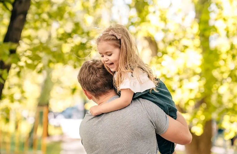 Man carrying smiling young girl on his back outdoors with green trees in background.
