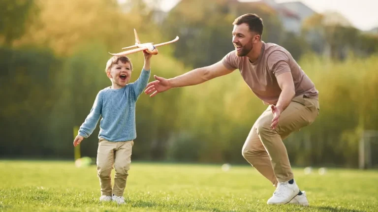 Young boy holding a toy airplane, smiling, with an adult man reaching out to him in a grassy park.