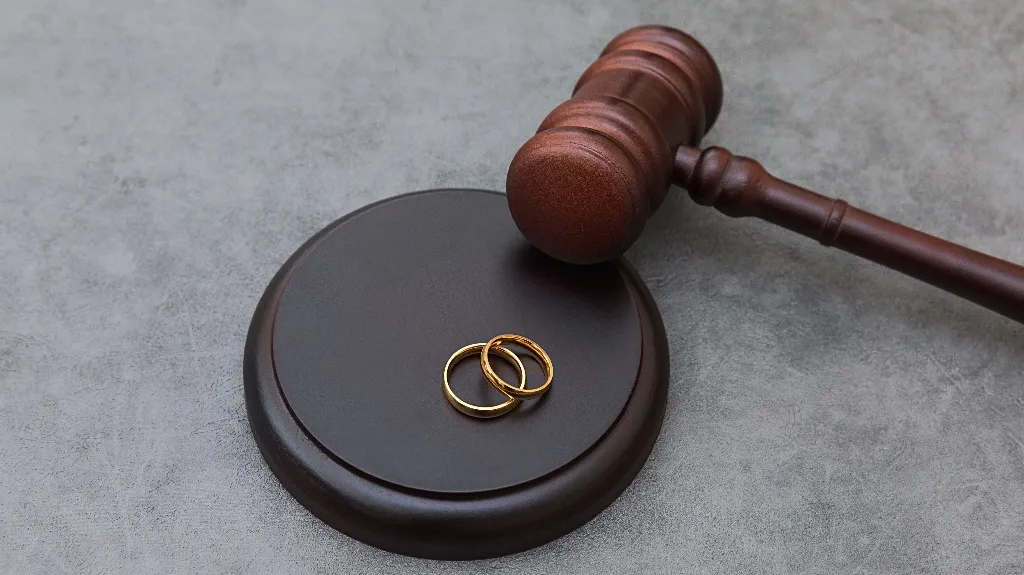 Wooden judge's gavel resting on a dark circular base with two intertwined gold wedding rings.