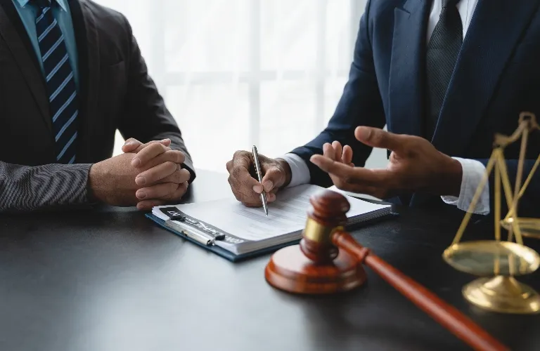 Two men in suits discussing legal documents with a gavel and scales of justice on the table.