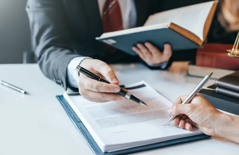 Two professionals in suits reviewing and signing documents at a white desk.
