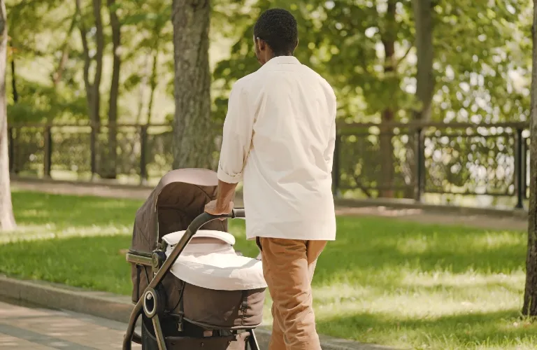 Adult male pushing a brown baby stroller along a park pathway with green trees and grass.