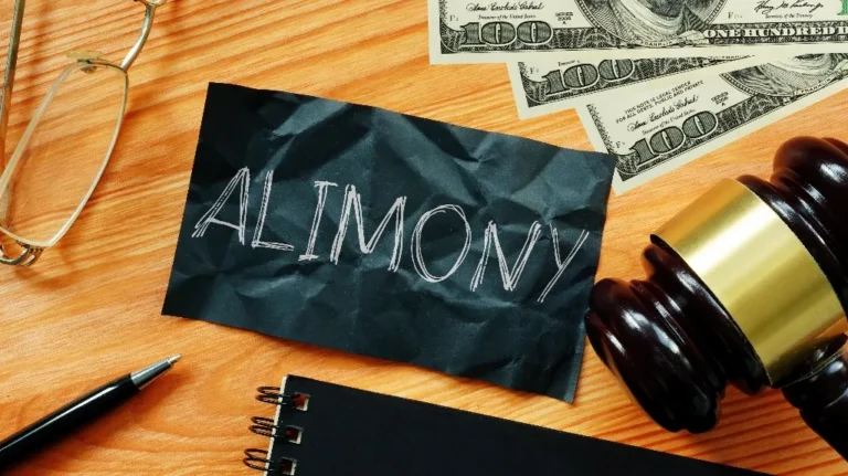 Black paper with "ALIMONY" text, wooden gavel, US hundred-dollar bills, glasses, pen, and notebook on wooden surface.