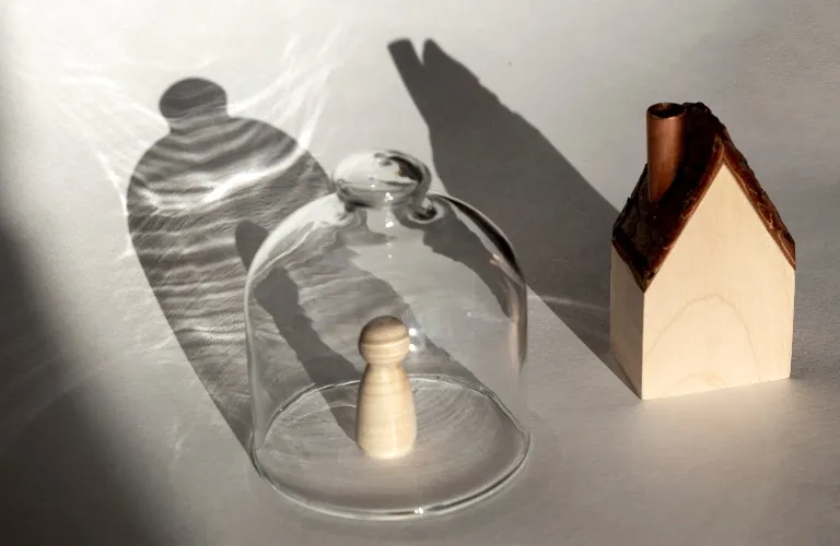 Wooden pawn figure under a glass dome casting a shadow, next to a small wooden house model.