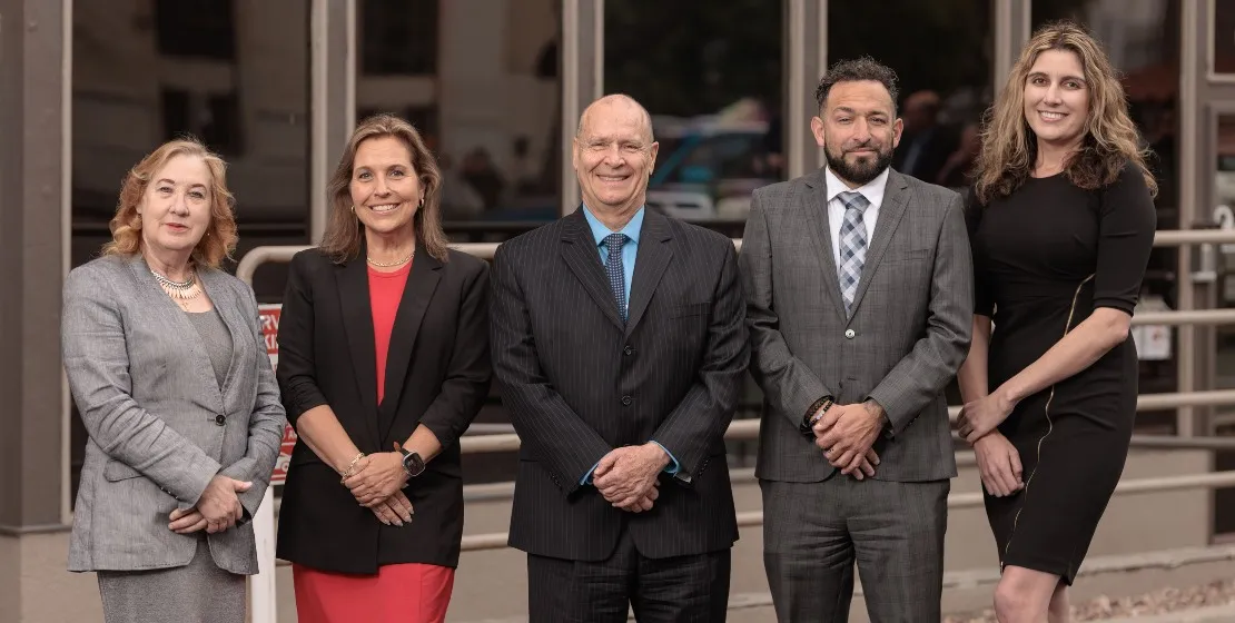 Group of five professionally dressed individuals standing outdoors, smiling at camera.