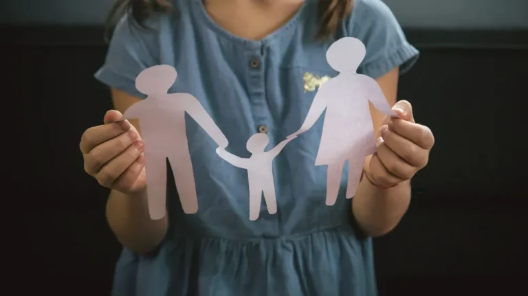 Child wearing blue dress holding paper cutout of family figures against dark background.