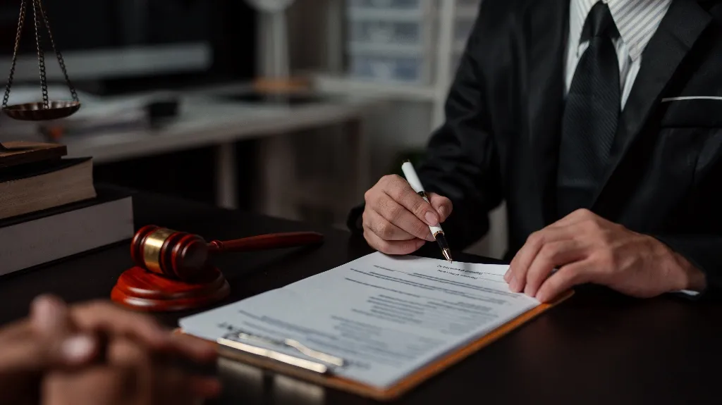 Person in dark suit signing legal document on desk with gavel and scales of justice.