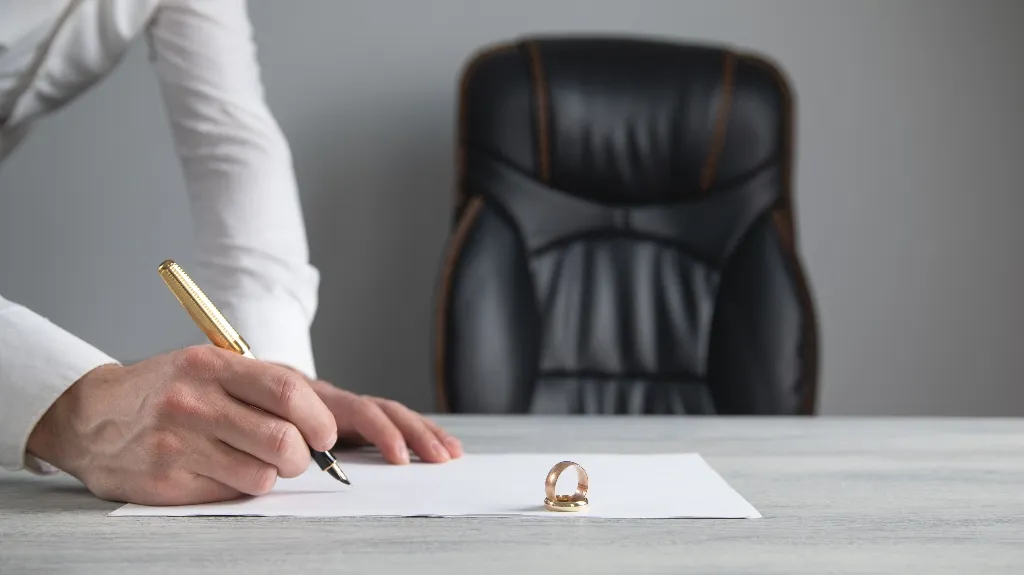 Person in white shirt signing document on desk with gold wedding ring placed on paper.