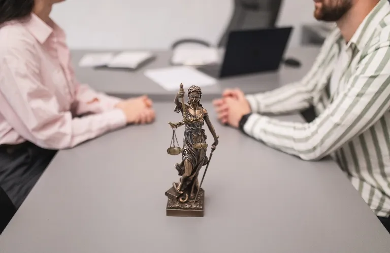 Bronze Lady Justice statue on a gray table between two seated individuals in a professional setting.