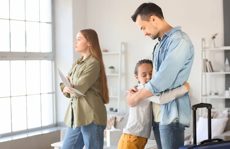 A woman stands looking out a window holding papers, while a man embraces a child in a living room.