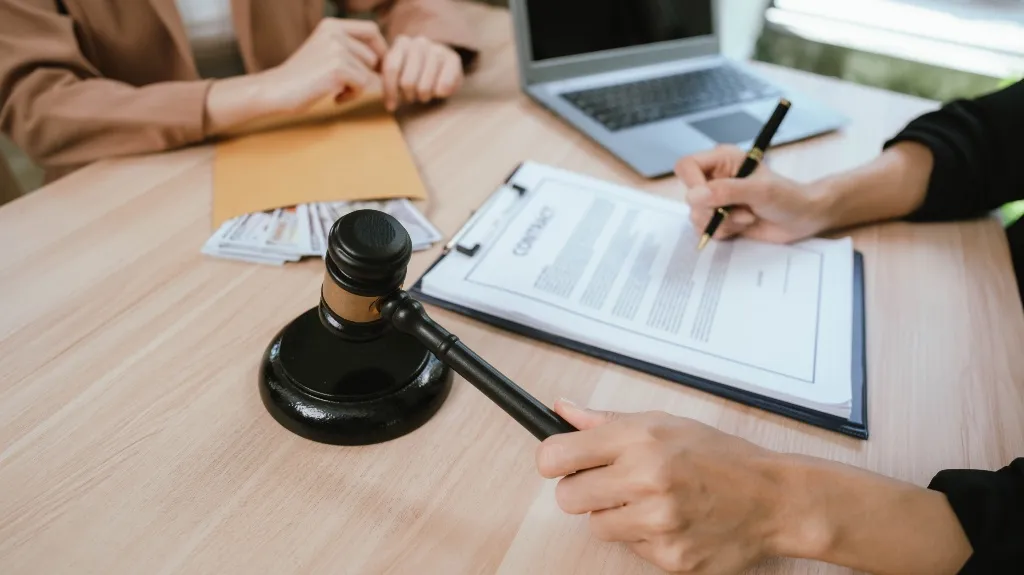Two individuals at a desk with a contract, money, laptop, and a judge's gavel.