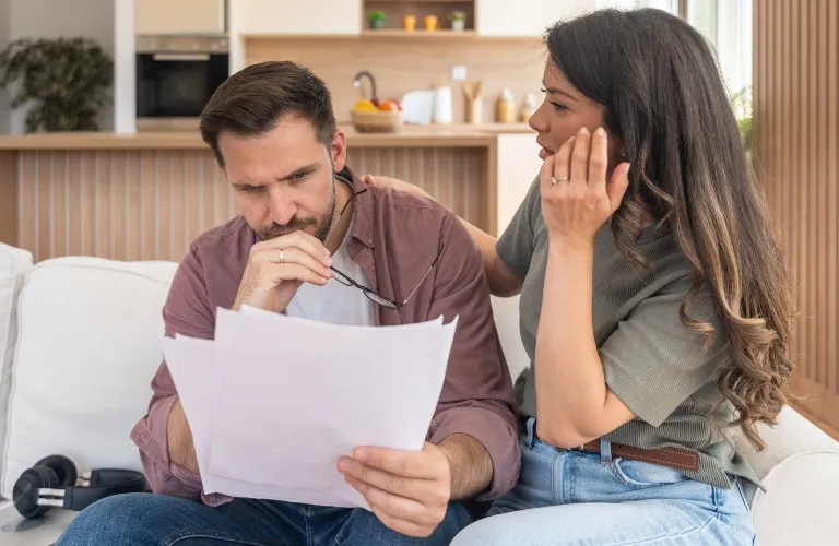 A man intently reviewing documents while a woman beside him gestures, seated in a modern living room.