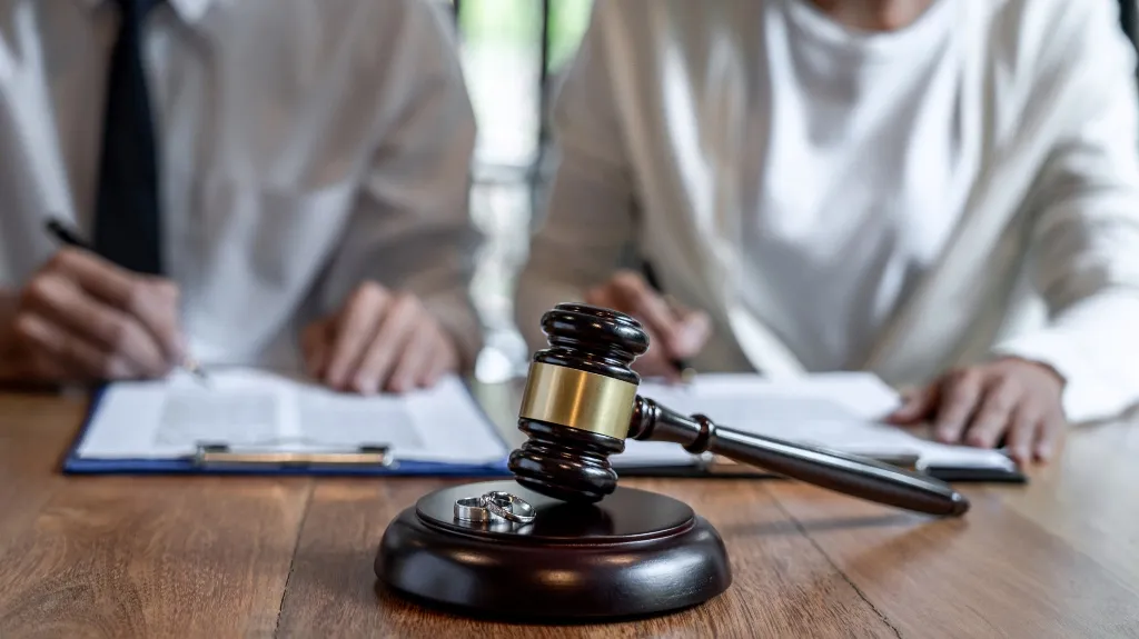 Wooden judge's gavel with two wedding rings on a table, blurred couple signing documents in background.