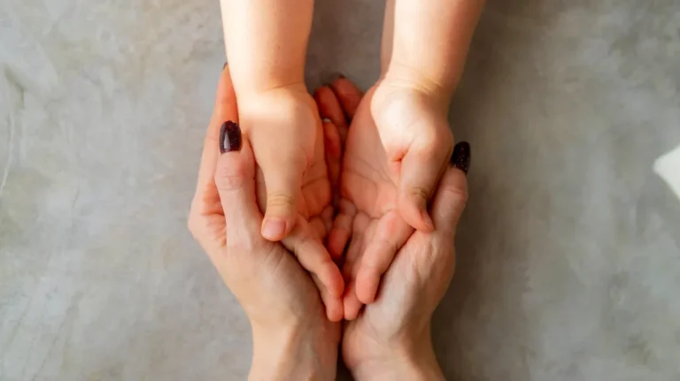 Adult hands gently holding a child's cupped hands against a gray background.