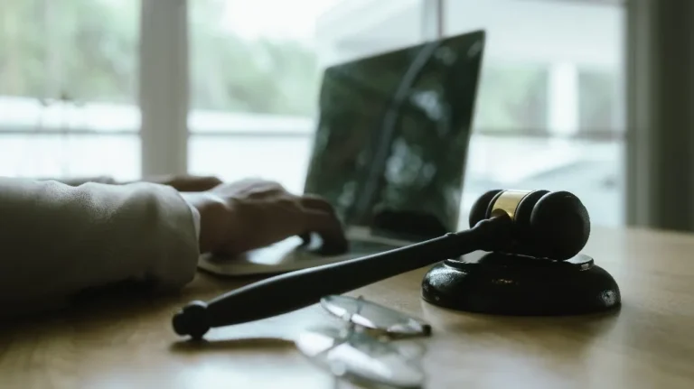 Wooden judge's gavel on table with blurred laptop and person typing in background.