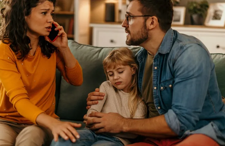 A concerned woman talks on a phone while a man comforts a sad young girl on a couch.