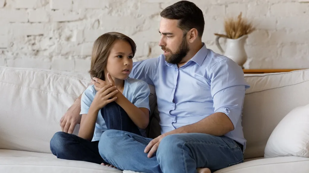 Man in blue shirt attentively talking with young boy on white couch.
