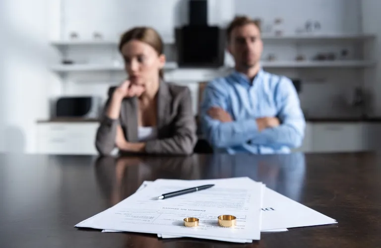 Two individuals sitting with arms crossed behind divorce papers, pen, and wedding rings on table.