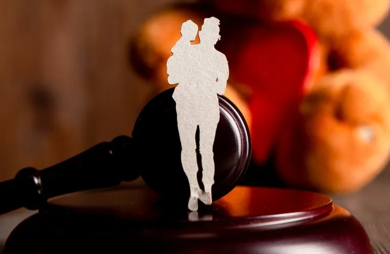 Silhouette cutout of a person holding a child placed on a wooden judge's gavel with blurred background.