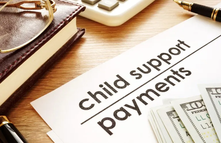 Document titled "child support payments" on wooden desk with pen, calculator, glasses, book, and US currency.