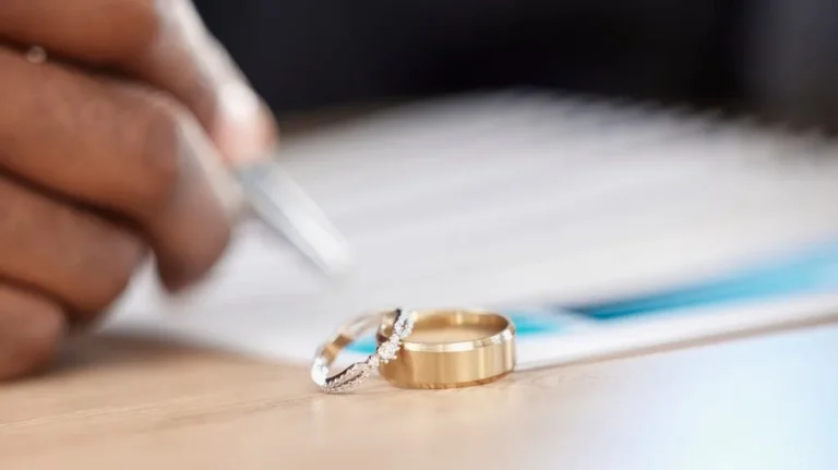 Close-up of two wedding rings on a wooden surface with a hand signing a document in the background.