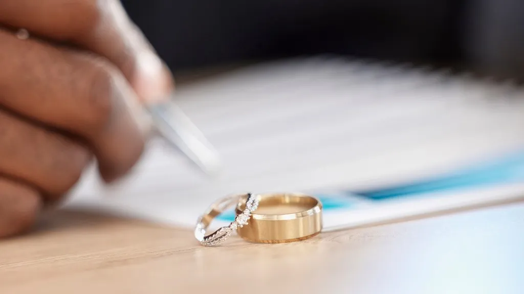 Close-up of two wedding rings on a wooden surface with a hand signing a document in the background.