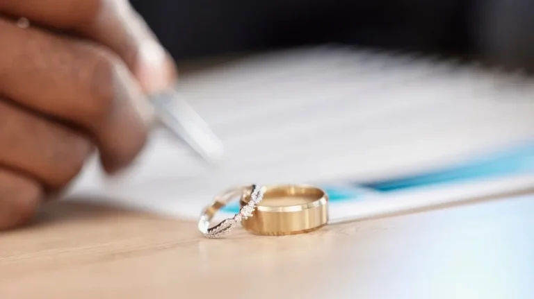 Close-up of two wedding rings on a table with a hand holding a pen over a document.