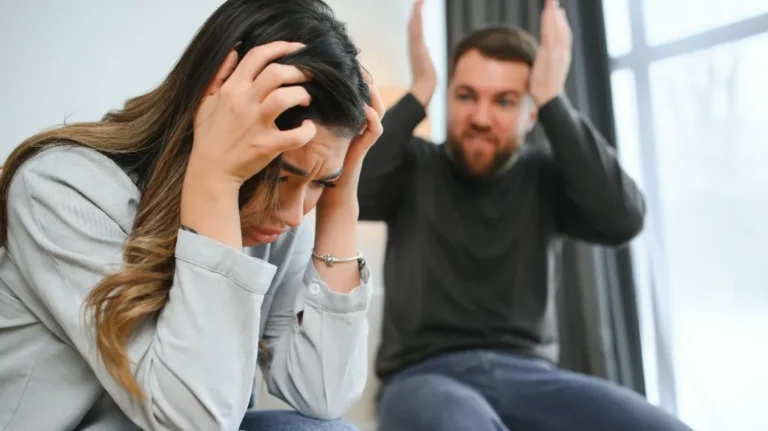 Frustrated woman holding her head with distressed man gesturing in the background indoors.