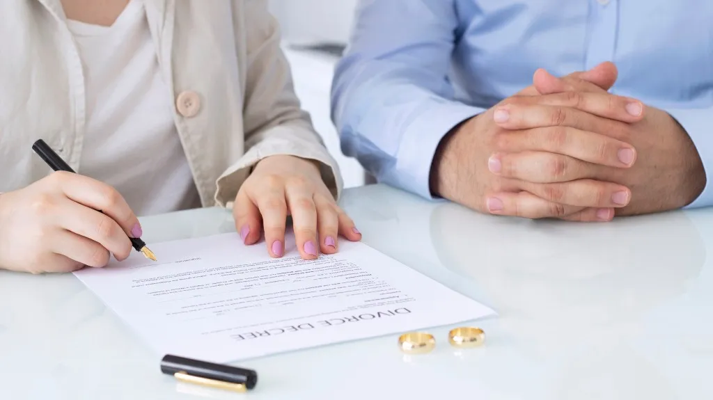 Two individuals at a table, one signing a divorce decree document, with two gold rings placed nearby.