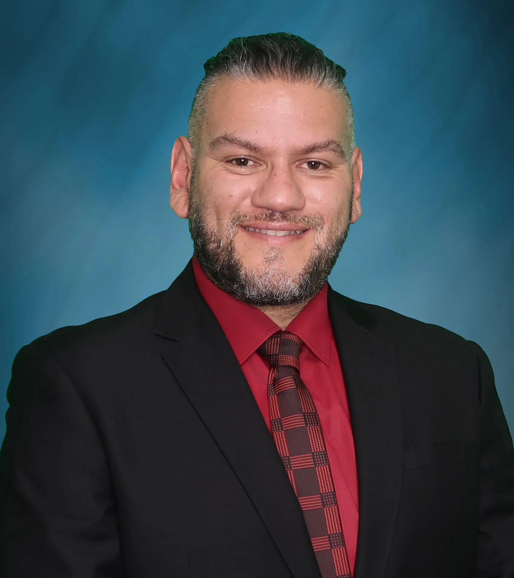 Professional headshot of a smiling man with gray-streaked hair and beard, wearing a black suit, red shirt, and patterned tie.