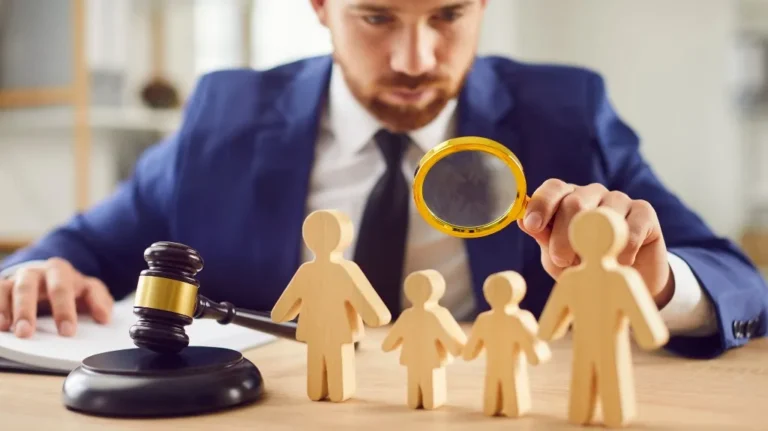 Man in blue suit examining wooden family figures with magnifying glass, gavel on desk.